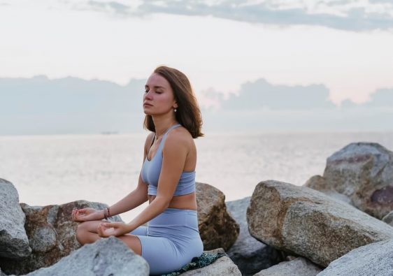 Woman in activewear meditating peacefully on rocks by the ocean at sunrise, promoting wellness and tranquility.