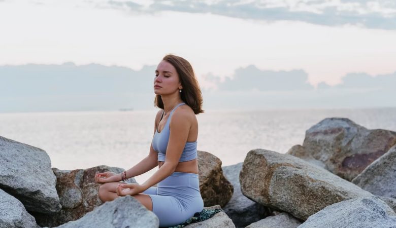 Woman in activewear meditating peacefully on rocks by the ocean at sunrise, promoting wellness and tranquility.