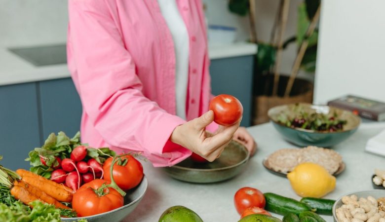 Person in pink overshirt holding a fresh tomato with various organic vegetables on a countertop.